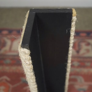 Op de afbeelding: Close-up van een zwart houten plank met een touwrand. De plank is aan een muur gemonteerd en heeft een eenvoudig, modern ontwerp.
