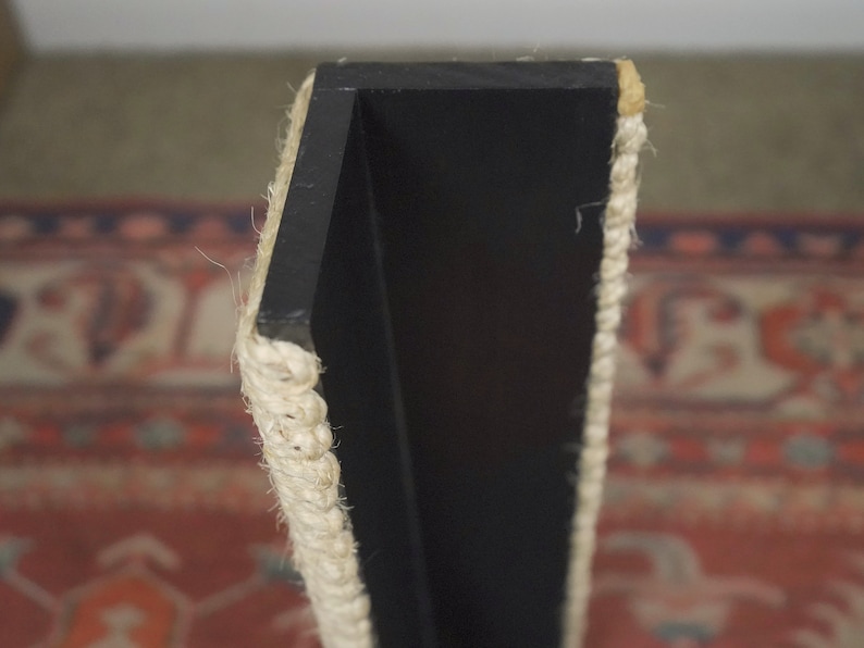Op de afbeelding: Close-up van een zwart houten plank met een touwrand. De plank is aan een muur gemonteerd en heeft een eenvoudig, modern ontwerp.