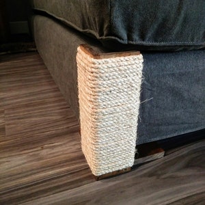 Op de afbeelding: Close-up van een grijze bank met een met touw omwikkelde houten poot. Het touw is wit en strak om de poot gewikkeld, die vierkant is. De bank staat op een houten vloer.