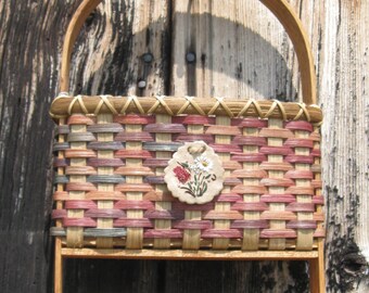 May include: A woven basket with a floral design hangs on a wooden wall with three hooks. The basket is made of natural materials and has a light brown, pink, and gray color scheme.