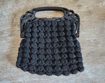 Puede incluir: Un bolso de mano de crochet negro con un asa de plástico negro. El asa es ornamentada y presenta un diseño de pájaro.