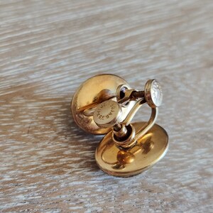 May include: A pair of gold-tone clip-on earrings with a round, slightly oval-shaped design. The earrings have a screw-back closure and are marked "MAR 14K GF".