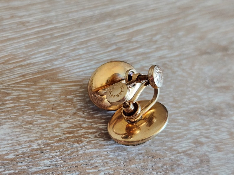 May include: A pair of gold-tone clip-on earrings with a round, slightly oval-shaped design. The earrings have a screw-back closure and are marked "MAR 14K GF".