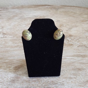May include: A pair of green and gold oval shaped earrings with a scarab beetle design on a black display stand.