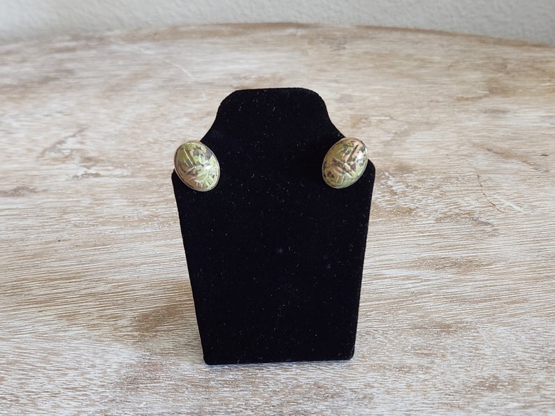 May include: A pair of green and gold oval shaped earrings with a scarab beetle design on a black display stand.