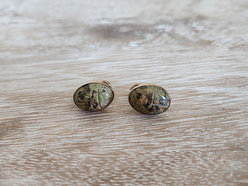 May include: A pair of gold tone earrings with green and brown oval shaped stones. The stones have a marbled pattern.