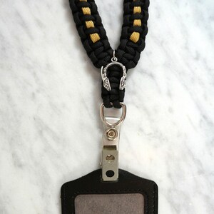 May include: Black and gold braided lanyard with a silver headphone charm and a black ID badge holder. The ID holder has a clear window and a metal clip. The text "RainyDayzArt" is at the bottom.