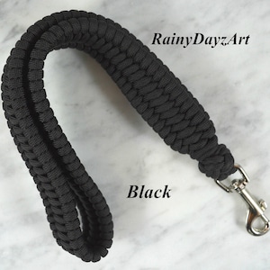 May include: A black paracord wrist strap featuring a braided design and a silver-toned metal swivel lobster clasp. The text RainyDayzArt and Black are visible on the image. The strap is formed into a loop, showcasing the textured cord construction.