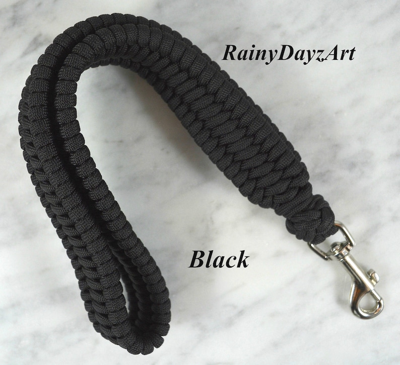 May include: A black paracord wrist strap featuring a braided design and a silver-toned metal swivel lobster clasp. The text RainyDayzArt and Black are visible on the image. The strap is formed into a loop, showcasing the textured cord construction.
