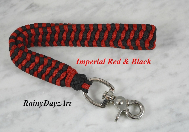 May include: A wrist lanyard crafted from black and red paracord in a braided pattern, featuring a silver-toned metal swivel lobster clasp. The text Imperial Red & Black and RainyDayzArt is printed in red and black on the surface.