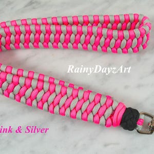 May include: A wrist lanyard crafted from neon pink and silver paracord in a braided pattern, featuring a black knot detail and a silver-toned metal swivel lobster clasp. The text RainyDayzArt and Neon Pink & Silver is visible on the image.