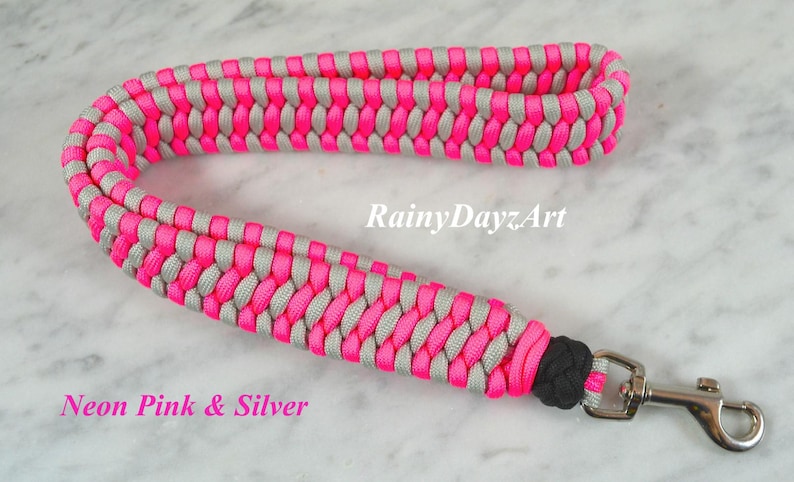 May include: A wrist lanyard crafted from neon pink and silver paracord in a braided pattern, featuring a black knot detail and a silver-toned metal swivel lobster clasp. The text RainyDayzArt and Neon Pink & Silver is visible on the image.