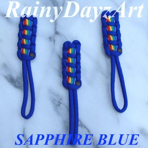 May include: Three zipper pulls made of sapphire blue paracord featuring a central rainbow-colored woven pattern. Each pull has a looped cord at the bottom and a small dark circular accent. The text RainyDayzArt and SAPPHIRE BLUE is visible.