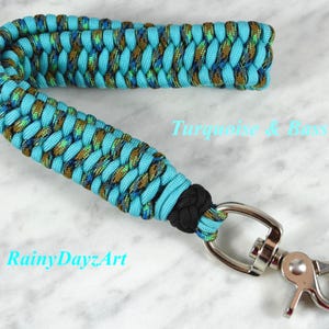 May include: A wrist lanyard crafted from braided paracord in turquoise and a metallic bass-colored pattern. The strap features a black knot near the base and is attached to a silver-toned metal swivel lobster clasp. Text reads Turquoise & Bass.