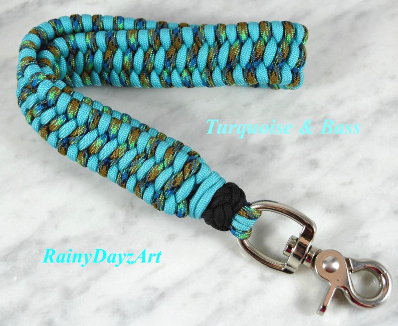 May include: A wrist lanyard crafted from braided paracord in turquoise and a metallic bass-colored pattern. The strap features a black knot near the base and is attached to a silver-toned metal swivel lobster clasp. Text reads Turquoise & Bass.
