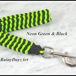 May include: A paracord leash handle featuring a braided pattern in neon green and black. The handle is attached to a silver-colored metal swivel snap hook. The text "Neon Green & Black" and "RainyDayzArt" is printed on the image.