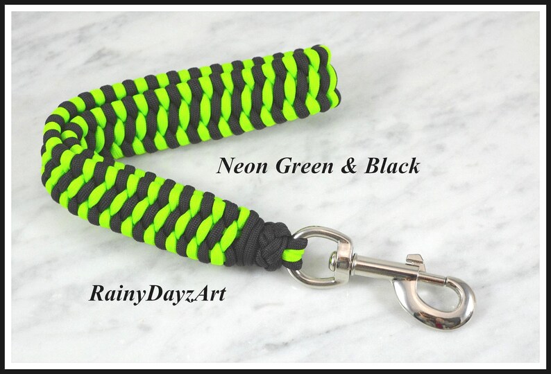 May include: A paracord leash handle featuring a braided pattern in neon green and black. The handle is attached to a silver-colored metal swivel snap hook. The text "Neon Green & Black" and "RainyDayzArt" is printed on the image.
