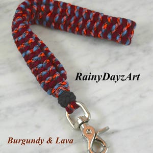 May include: A wrist lanyard crafted from braided paracord in a burgundy, light blue, and bright orange pattern. The cord is secured with a black knot and features a silver-toned metal swivel lobster clasp. The text RainyDayzArt and Burgundy & Lava appear.