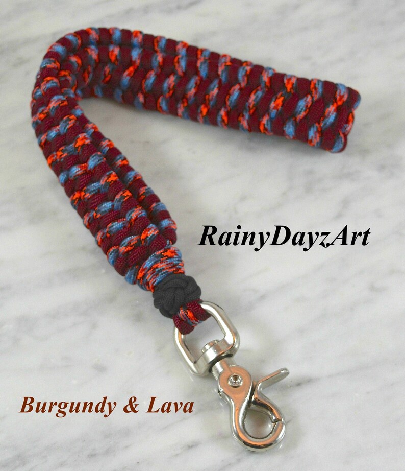 May include: A wrist lanyard crafted from braided paracord in a burgundy, light blue, and bright orange pattern. The cord is secured with a black knot and features a silver-toned metal swivel lobster clasp. The text RainyDayzArt and Burgundy & Lava appear.