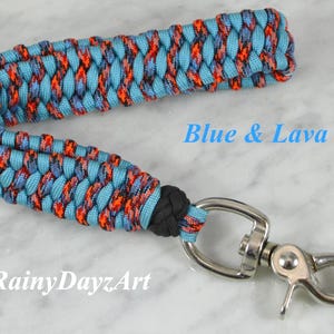 May include: A wrist lanyard made of braided paracord in light blue and a multi-colored pattern of orange, black, and blue. It features a black knot and a silver-toned metal swivel lobster clasp. The text Blue & Lava 2 and RainyDayzArt is visible.