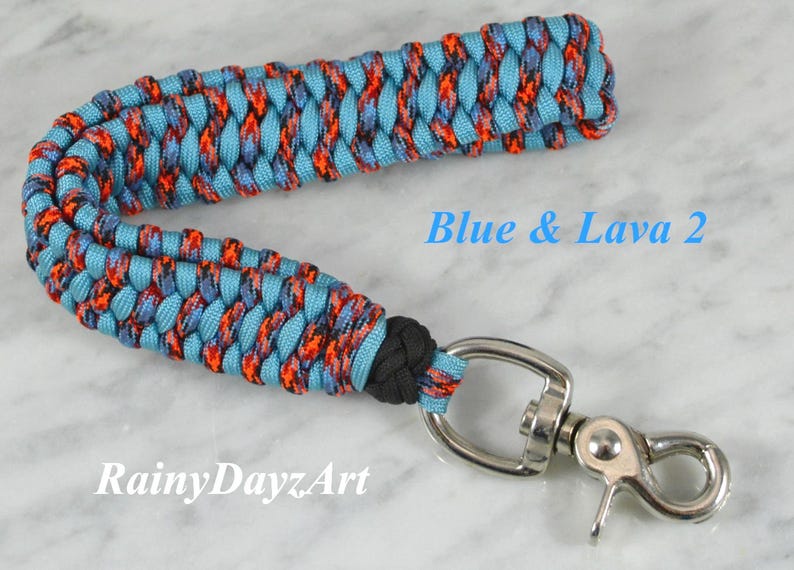 May include: A wrist lanyard made of braided paracord in light blue and a multi-colored pattern of orange, black, and blue. It features a black knot and a silver-toned metal swivel lobster clasp. The text Blue & Lava 2 and RainyDayzArt is visible.