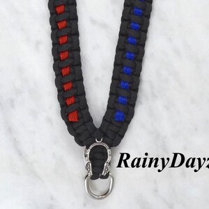 May include: Black lanyard with red and blue accents. The lanyard is made of woven material and features a silver-colored metal clasp and ring. The text "RainyDayzArt" is visible in the image.