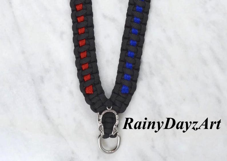 May include: Black lanyard with red and blue accents. The lanyard is made of woven material and features a silver-colored metal clasp and ring. The text "RainyDayzArt" is visible in the image.