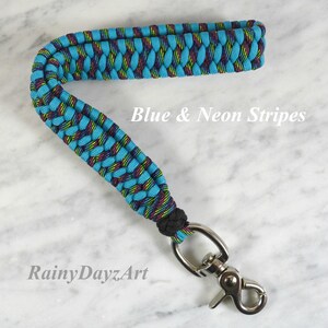 May include: A wrist lanyard made of braided paracord in bright blue and neon striped patterns. The strap features a black knot at the base and a silver-toned metal swivel lobster clasp. The text Blue & Neon Stripes and RainyDayzArt is visible.