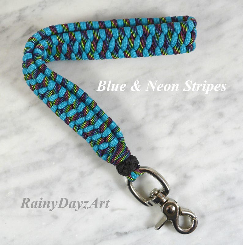 May include: A wrist lanyard made of braided paracord in bright blue and neon striped patterns. The strap features a black knot at the base and a silver-toned metal swivel lobster clasp. The text Blue & Neon Stripes and RainyDayzArt is visible.