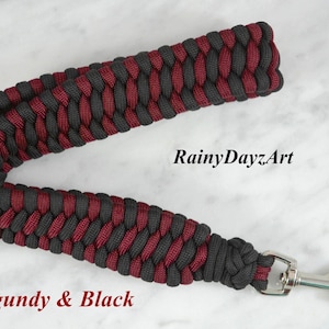 May include: A paracord wrist lanyard featuring a braided pattern in burgundy and black. The strap is attached to a silver-colored metal swivel snap hook. The text RainyDayzArt and Burgundy & Black are visible on the image.