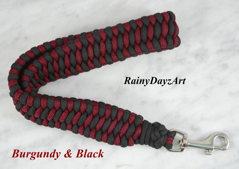 May include: A paracord wrist lanyard featuring a braided pattern in burgundy and black. The strap is attached to a silver-colored metal swivel snap hook. The text RainyDayzArt and Burgundy & Black are visible on the image.