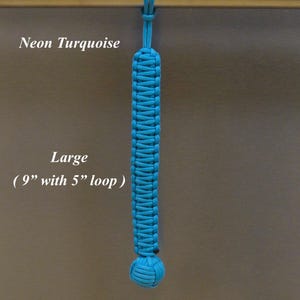 May include: A neon turquoise paracord lanyard featuring a braided rectangular body and a spherical knot at the bottom. The text Neon Turquoise and Large ( 9" with 5" loop ) and RainyDayzArt are visible. The lanyard hangs from a horizontal bar.