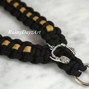 May include: Black and gold paracord dog collar with a silver-colored headphone charm and a metal ring. The collar is made of woven black and gold cord. The text "RainyDayzArt" is visible.