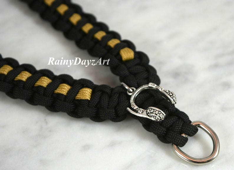 May include: Black and gold paracord dog collar with a silver-colored headphone charm and a metal ring. The collar is made of woven black and gold cord. The text "RainyDayzArt" is visible.
