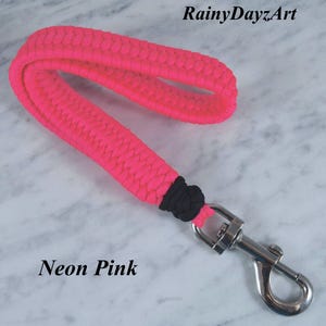 May include: A neon pink paracord wrist lanyard featuring a braided design, a black knot accent, and a silver-toned metal swivel lobster clasp. The text RainyDayzArt and Neon Pink are visible in the image.