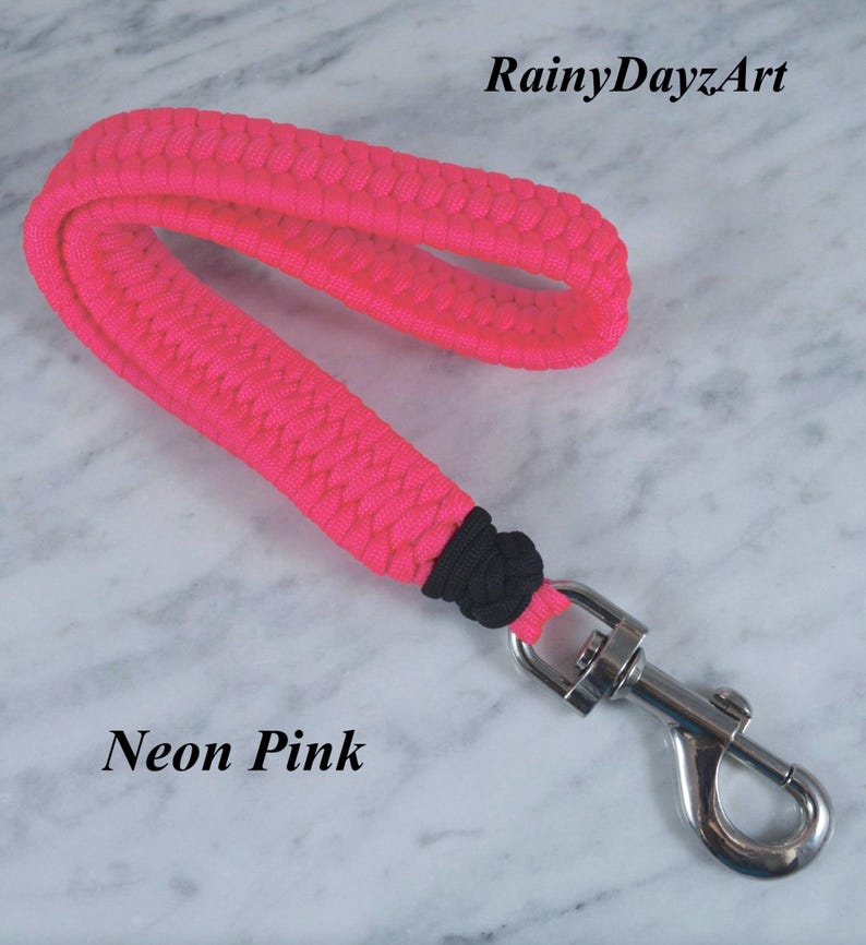 May include: A neon pink paracord wrist lanyard featuring a braided design, a black knot accent, and a silver-toned metal swivel lobster clasp. The text RainyDayzArt and Neon Pink are visible in the image.