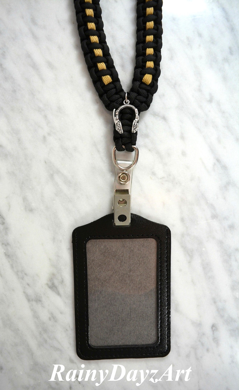 May include: Black and gold braided lanyard with a silver headphone charm and a black ID badge holder. The ID holder has a clear window and a metal clip. The text "RainyDayzArt" is at the bottom.