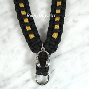May include: Black and gold braided lanyard with a silver headphone charm and a silver ring at the bottom. The lanyard has the text "RainyDayzArt" in the upper center.