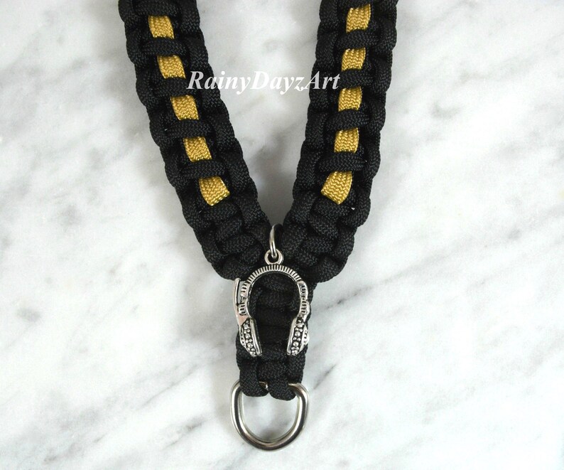 May include: Black and gold braided lanyard with a silver headphone charm and a silver ring at the bottom. The lanyard has the text "RainyDayzArt" in the upper center.