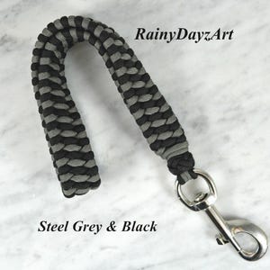 May include: A wrist lanyard crafted from black and steel grey paracord in a braided pattern, featuring a silver-toned metal swivel snap hook. The text RainyDayzArt and Steel Grey & Black are visible in the image.