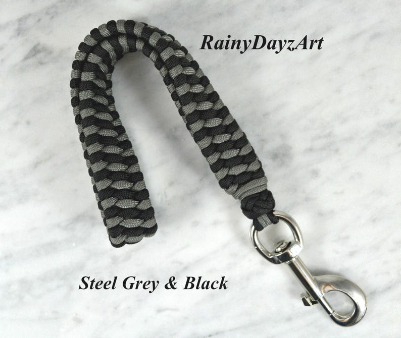 May include: A wrist lanyard crafted from black and steel grey paracord in a braided pattern, featuring a silver-toned metal swivel snap hook. The text RainyDayzArt and Steel Grey & Black are visible in the image.