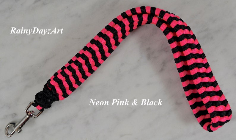 May include: A wrist lanyard crafted from neon pink and black paracord in a braided pattern, featuring a silver-toned metal swivel clasp. The text RainyDayzArt and Neon Pink & Black is visible on the image.