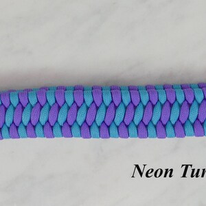 May include: A wrist lanyard made of braided neon turquoise and lilac paracord, featuring a silver-toned metal swivel lobster clasp at one end. The text RainyDayzArt and Neon Turquoise & Lilac are printed on the image near the braided cord.