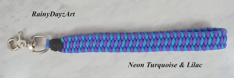 May include: A wrist lanyard made of braided neon turquoise and lilac paracord, featuring a silver-toned metal swivel lobster clasp at one end. The text RainyDayzArt and Neon Turquoise & Lilac are printed on the image near the braided cord.