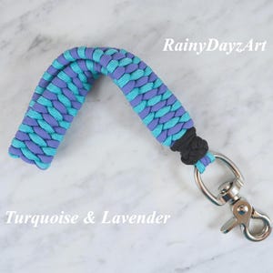 May include: A paracord keychain featuring a braided pattern in turquoise and lavender colors. The strap is secured with a black knot and attached to a silver-colored metal swivel lobster clasp. The text RainyDayzArt and Turquoise & Lavender appear.