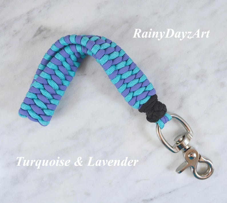 May include: A paracord keychain featuring a braided pattern in turquoise and lavender colors. The strap is secured with a black knot and attached to a silver-colored metal swivel lobster clasp. The text RainyDayzArt and Turquoise & Lavender appear.