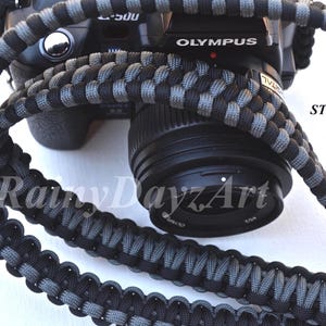 May include: A camera strap woven in a black and steel grey paracord pattern, draped over a black Olympus camera. The text "BLACK & STEEL GREY" and "RainyDayzArt" are visible on the image, indicating the color scheme and the brand name.