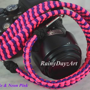 May include: A camera strap woven from neon pink and dark purple cord in a thick, textured pattern. The strap features black cord knots at the attachment points and is draped over a black digital camera. The text RainyDayzArt is visible.
