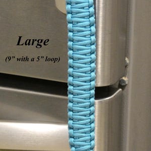 May include: A large paracord lanyard featuring a light blue braided handle and a black monkey fist knot at the end. The lanyard includes a black loop and measures 9 inches in total length with a 5 inch loop. Text on the image reads Blue & Black, Large, (9” with a 5” loop), and RainyDayzArt.