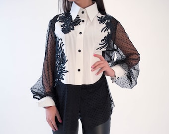 May include: A white button-down shirt with black floral embroidery on the sleeves. The shirt has a pleated front and a collar. The model is wearing black leggings and black heels.
